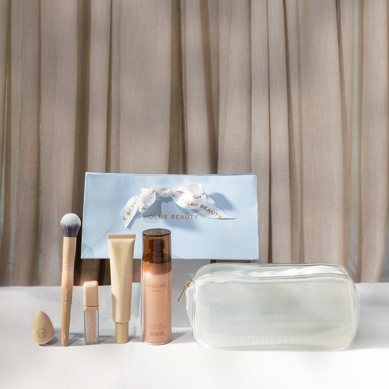 Makeup products including brushes and bottles on a white surface with a neutral curtain background